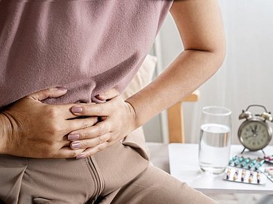 Frau hält sich den Bauch, im Hintergrund Medikamente.