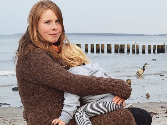 Blonde Frau sitzt mit Kleinkind im Arm am Strand.